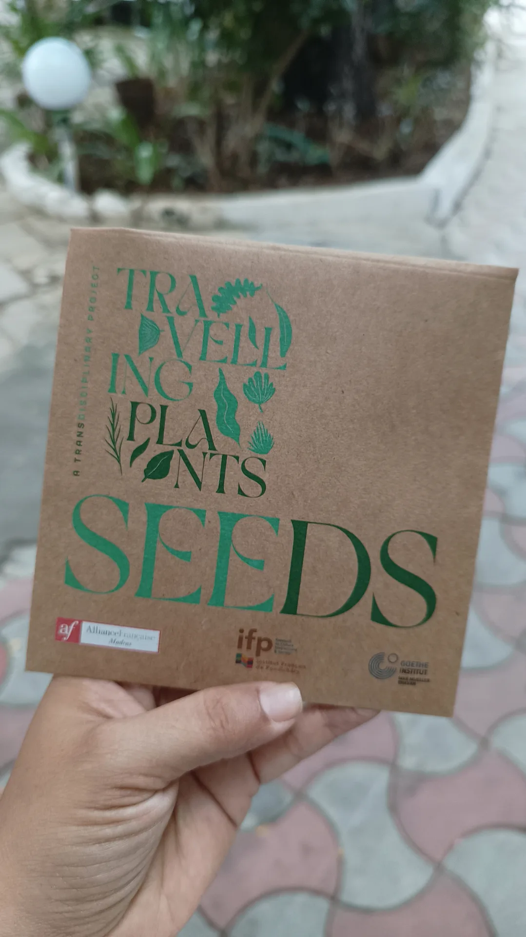 Hand holding Travelling Plants seeds package showcasing eco-friendly botanical project branding and transdisciplinary approach