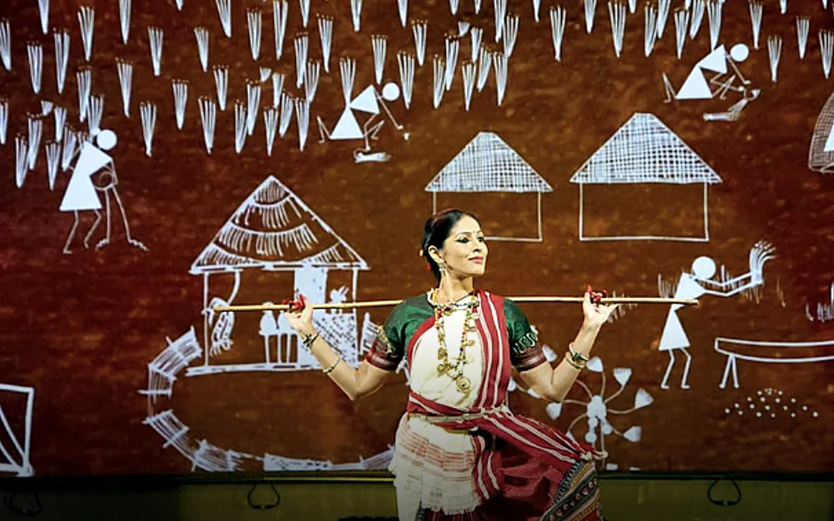 Bharatanatyam dancer holding a bamboo stick across her shoulders in front of a Warli art backdrop of huts, trees and figures — When Walls Dance